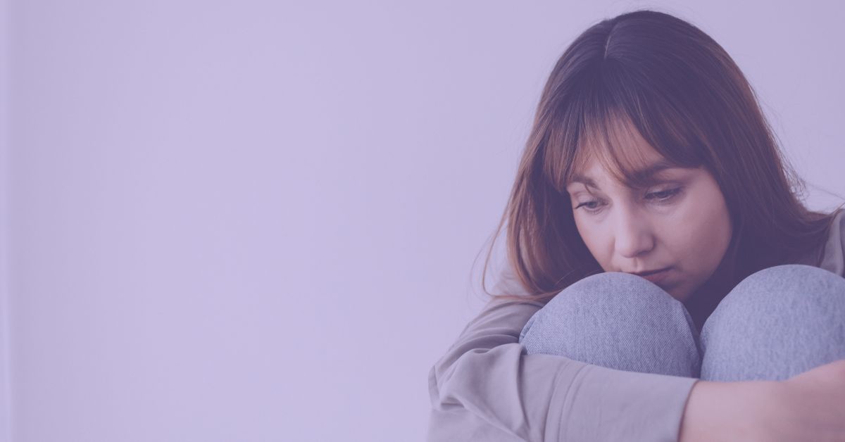 A young woman with brown hair hugs her knees tightly, head bowed in sadness against a purple backdrop, symbolising the emotional toll of fibromyalgia and depression.