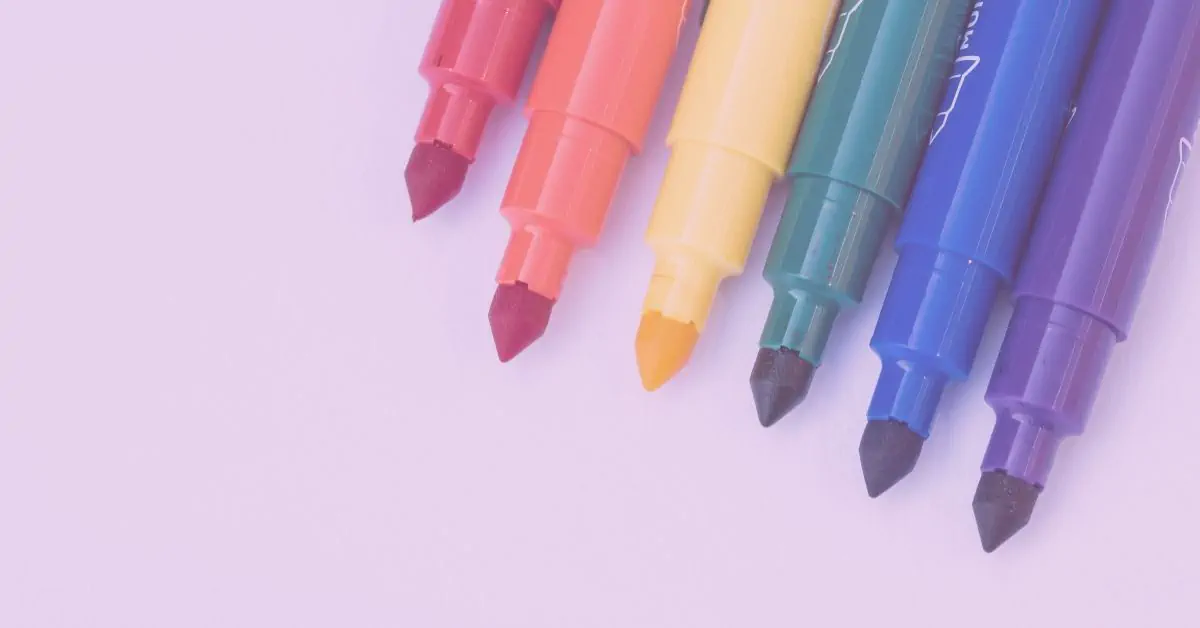 Row of colourful felt-tip markers in red, orange, yellow, green, blue and purple against a pale pink background – representing the vibrant, diverse essence of creativity and self-expression.