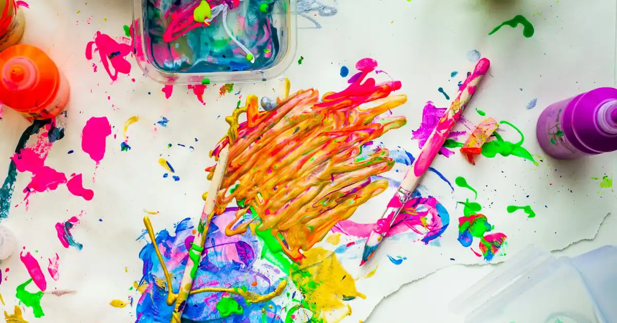Colourful paint swirls in yellow, pink, blue & green with brushes & bottles on white surface, showing creative mess.