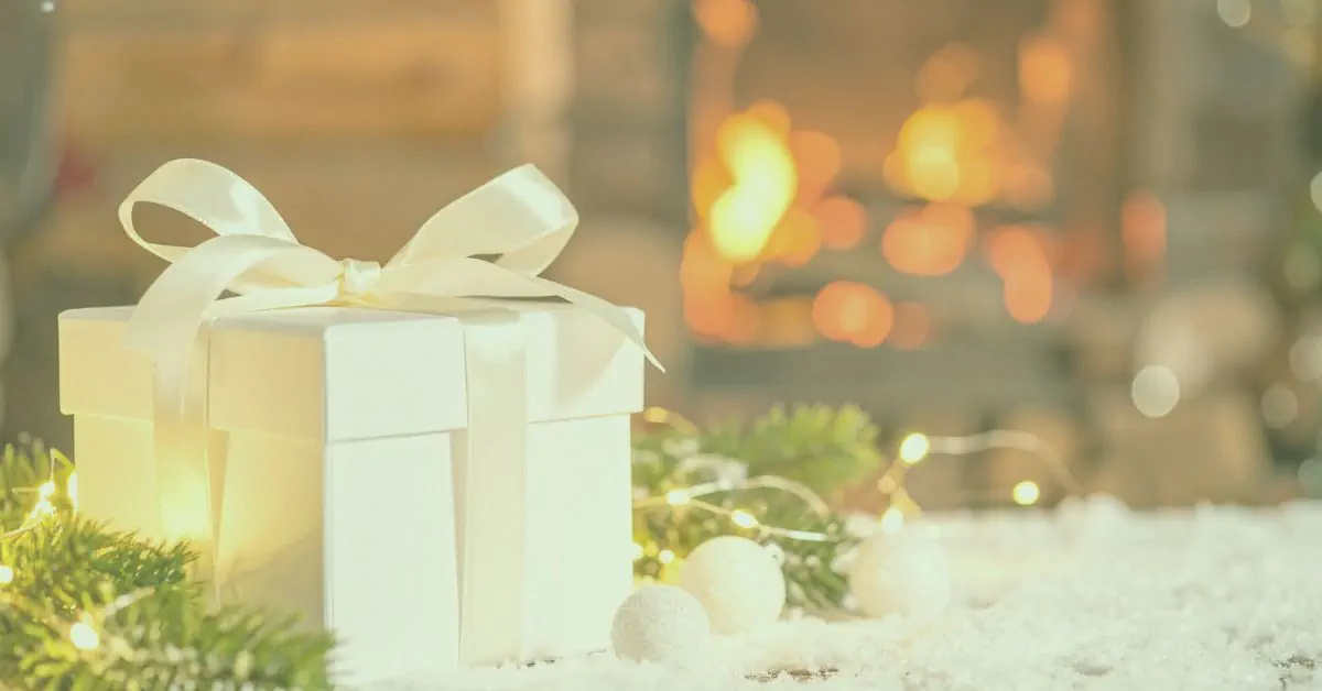 Elegant white Christmas gift box tied with cream satin ribbon, surrounded by fairy lights, pine branches and faux snow with a warm glowing fireplace in the background.