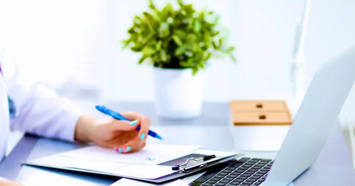 Person with Fibromyalgia writing on clipboard at desk with laptop and plant, illustrating adaptable working.